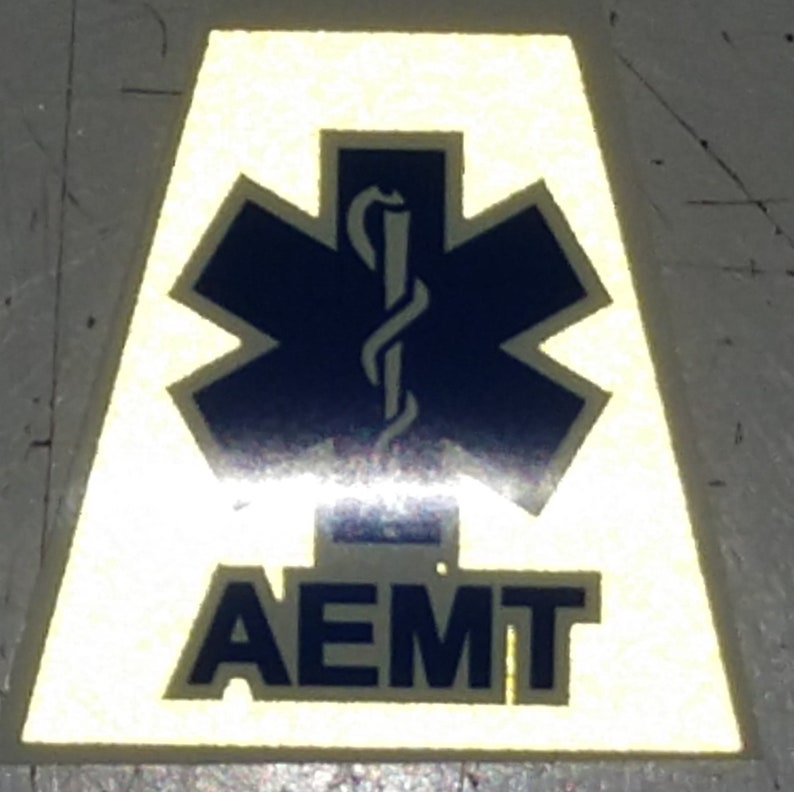 Pu&ograve; includere: Un adesivo riflettente giallo con una stella della vita blu e il testo "AEMT" in blu.