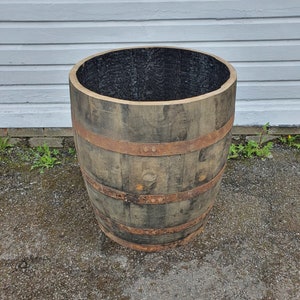 May include: A large wooden barrel planter with metal bands. The barrel is brown and has a dark brown interior. The barrel is sitting on a grey asphalt surface.