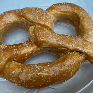 May include: A golden-brown pretzel sprinkled with coarse salt. The baked bread product is in a classic pretzel shape, with a glossy finish. The pretzel is on a clear, oval-shaped plate.