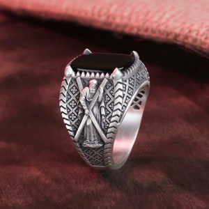 Peut inclure: Une bague en argent avec une pierre d'onyx noire et un motif complexe représentant une figure tenant des épées croisées.