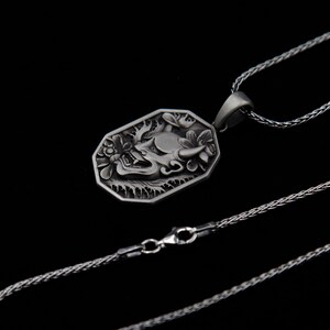 May include: A silver necklace with a detailed pendant featuring a stylized demon face. The pendant is octagonal and has a textured surface. The chain is a silver rope chain.