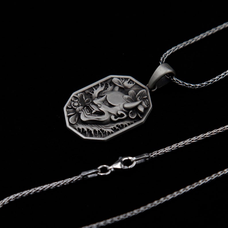 May include: A silver necklace with a detailed pendant featuring a stylized demon face. The pendant is octagonal and has a textured surface. The chain is a silver rope chain.
