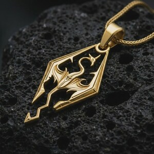 Gold plated Skyrim dragon pendant necklace on textured volcanic rock, premium close-up studio composition emphasizing the dragon symbol, geometric frame, and fantasy game-inspired design.