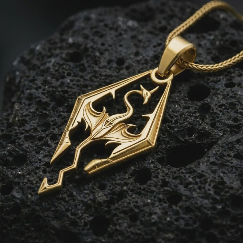 Gold plated Skyrim dragon pendant necklace on textured volcanic rock, premium close-up studio composition emphasizing the dragon symbol, geometric frame, and fantasy game-inspired design.