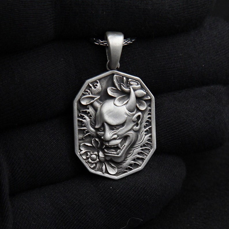 May include: Silver pendant necklace featuring a detailed, three-dimensional design of a Japanese oni demon mask with horns, surrounded by stylized floral and wave patterns.