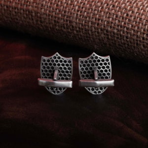 May include: A pair of silver cufflinks in the shape of a shield with a honeycomb pattern. The cufflinks are marked "925" on the back.
