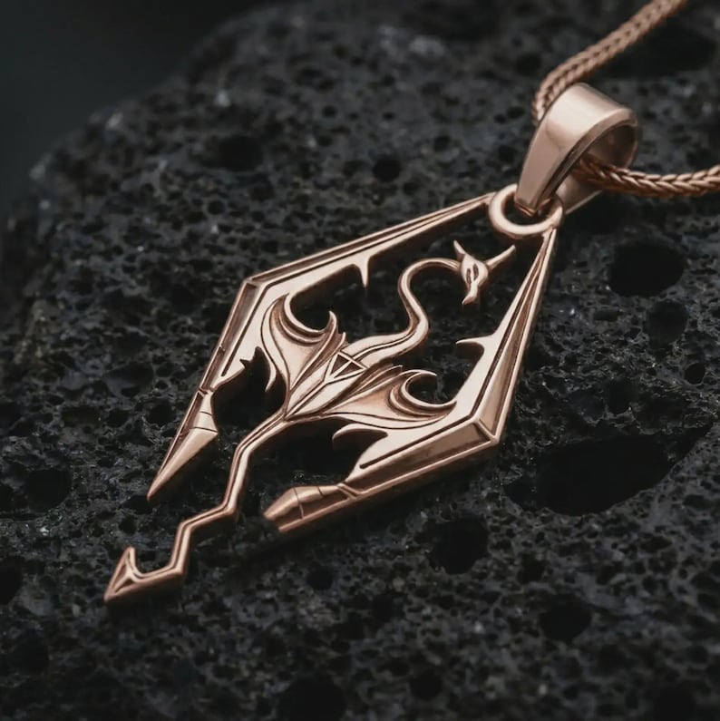 Rose gold plated Skyrim dragon pendant necklace on textured volcanic rock, detailed macro product shot with soft warm reflections and bold dragon emblem styling.