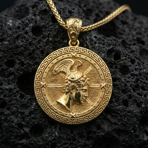 Gold plated Perseus coin pendant necklace on textured volcanic rock, premium close-up with warm reflective shine across the winged helmet, facial relief, and raised meander frame.