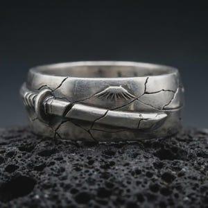Könnte beinhalten: Ein silberner Ring mit detailliertem Design. Der Ring hat eine rissige Textur und ein Katana-Schwert-Design. Der Ring hat auch ein Bergdesign. Der Ring ist ein einzigartiges Schmuckstück.