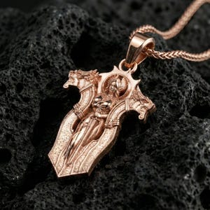 Rose gold plated gothic cross pendant necklace on textured volcanic rock, centered studio-style close-up with a soft rosy glow on the cross silhouette and sculpted figure detail.