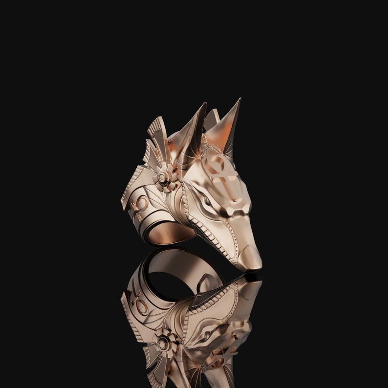 May include: A gold-toned ring with a detailed Anubis head design. The ring is set against a black background.