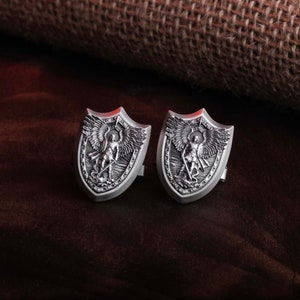 May include: A pair of silver cufflinks featuring a detailed design of an angel with wings, holding a sword and standing on a shield. The cufflinks are in the shape of a shield.
