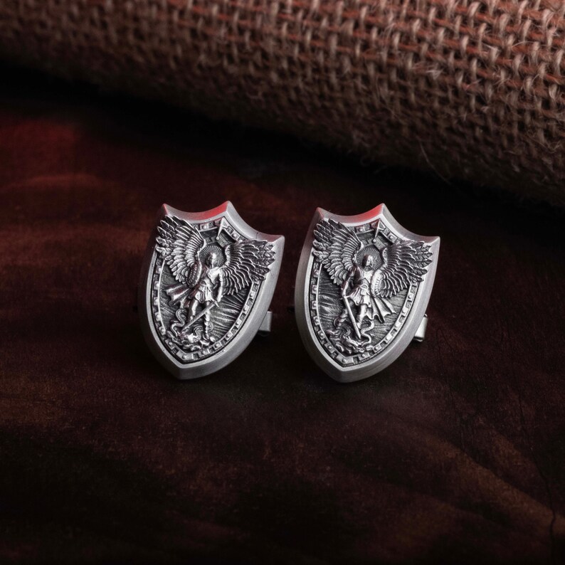 May include: A pair of silver cufflinks featuring a detailed design of an angel with wings, holding a sword and standing on a shield. The cufflinks are in the shape of a shield.