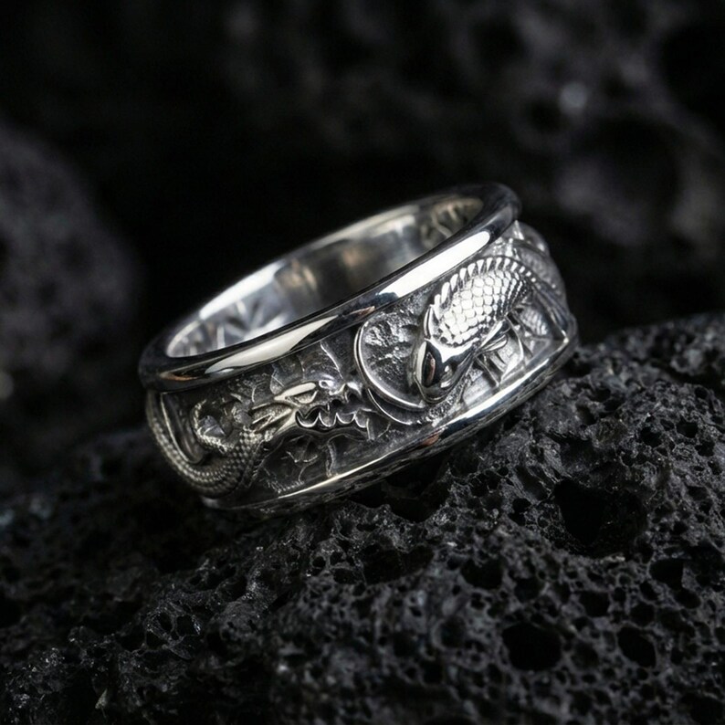 Polished silver dragon and koi fish wedding band on black volcanic stone, crisp sculpted relief with deep carved background, centered close-up studio lighting.