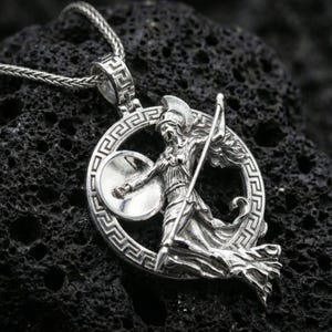Polished silver Athena warrior pendant necklace on textured volcanic rock, crisp macro product photo emphasizing clean edges, sharp relief detailing on the shield and spear, and bright reflective highlights.