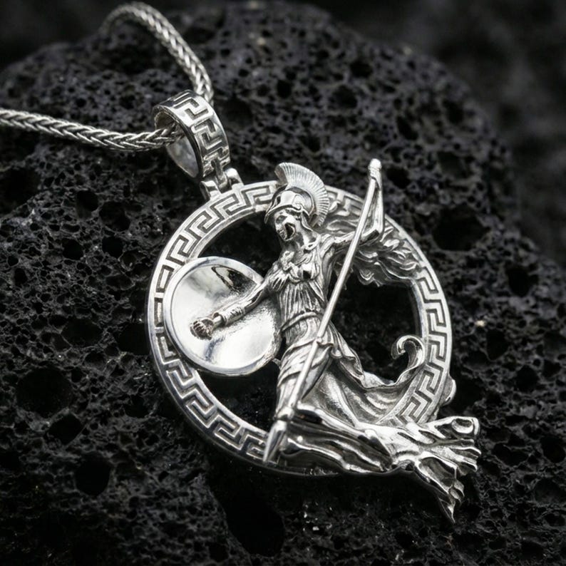 Polished silver Athena warrior pendant necklace on textured volcanic rock, crisp macro product photo emphasizing clean edges, sharp relief detailing on the shield and spear, and bright reflective highlights.