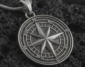 May include: A circular silver-toned pendant featuring an engraved compass rose design with cardinal directions N, NE, E, SE, S, SW, W, and NW marked along the outer rim. The pendant is attached to a silver-toned chain with a textured link pattern.
