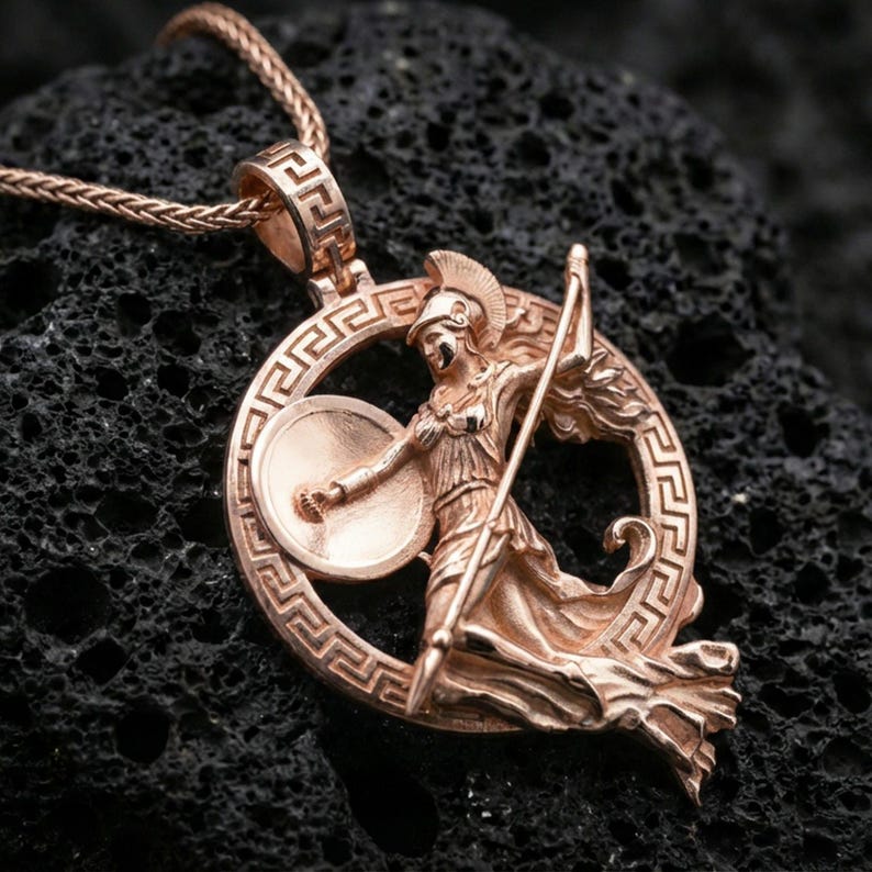 Rose gold plated Athena warrior pendant necklace on textured volcanic rock, centered studio-style close-up showcasing soft rosy glow and intricate Greek goddess armor-and-shield detailing.