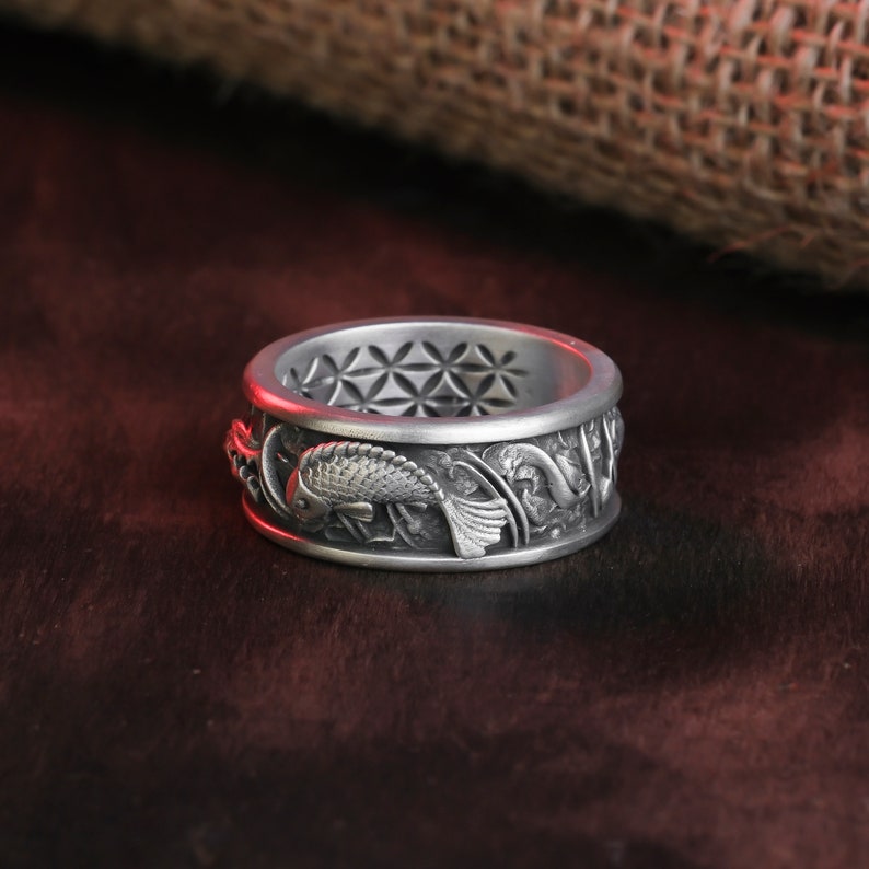 May include: A silver ring with a detailed koi fish design. The ring features a geometric pattern on the inner band and intricate aquatic plant and fish carvings around the outer band. The ring is displayed on a dark surface.