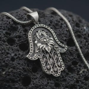 May include: A silver-toned Hamsa hand pendant necklace featuring an intricate embossed design of a central eye surrounded by floral-like petals and geometric patterns, suspended from a matching silver-toned chain with a polished metallic finish.