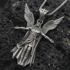 Op de afbeelding: Zilverkleurige hangerketting met een engel met uitgespreide vleugels en een kruis. De hanger, die aan een ketting hangt, toont een engel met uitgestrekte armen en een vloeiende cape. De ketting wordt gepresenteerd tegen een donkere, getextureerde achtergrond.