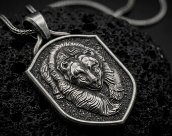 May include: A silver-toned pendant featuring a detailed relief of a bear head centered on a shield-shaped base with a textured background. The pendant is attached to a silver-toned chain, showcasing a polished finish on the bear and dark accents.