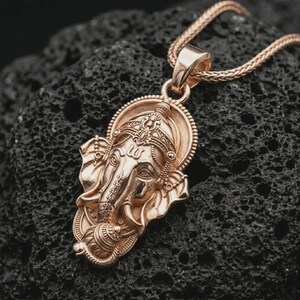 Rose gold plated Ganesha pendant necklace on textured volcanic rock, detailed macro product shot with soft warm reflections and sacred elephant deity design.