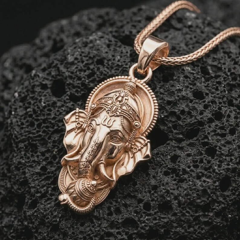Rose gold plated Ganesha pendant necklace on textured volcanic rock, detailed macro product shot with soft warm reflections and sacred elephant deity design.