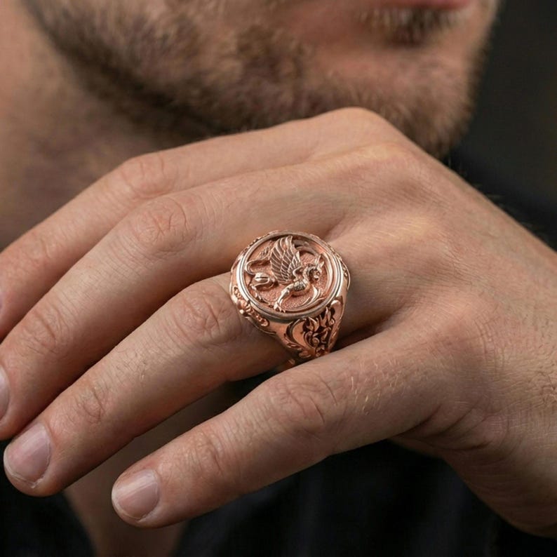 Rose gold plated griffon ring on finger featuring sculpted winged griffin emblem, refined fantasy jewelry lifestyle wear shot