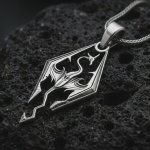 Polished silver Skyrim dragon pendant necklace on textured volcanic rock, centered macro jewelry photo with sharp fantasy emblem details and bright silver highlights.