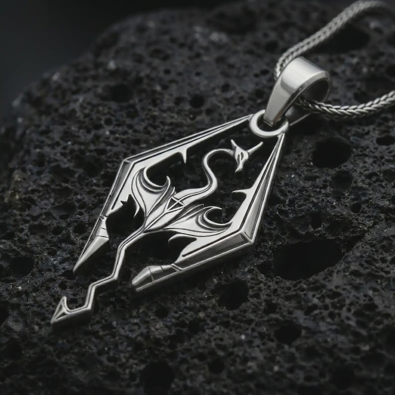 Polished silver Skyrim dragon pendant necklace on textured volcanic rock, centered macro jewelry photo with sharp fantasy emblem details and bright silver highlights.