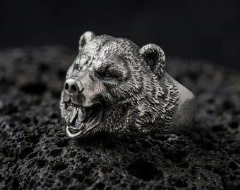 May include: A silver-toned metal ring featuring a highly detailed, three-dimensional bear head with an open mouth, visible teeth, and textured fur patterns. The metallic surface has a dark, oxidized finish that highlights the intricate carvings.
