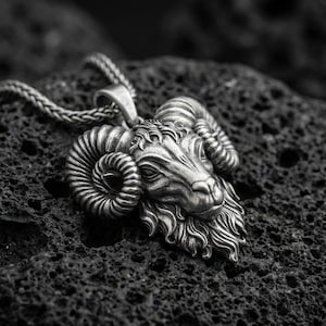 Op de afbeelding: Zilverkleurige ram-hanger ketting. De hanger heeft een gedetailleerde rammenkop met gekrulde hoorns en een getextureerd oppervlak. De ketting heeft een gevlochten ketting. De hanger en ketting liggen op een donker, getextureerd oppervlak.