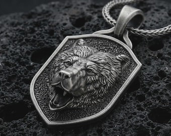May include: A silver-toned pendant necklace featuring a detailed bear head within a shield-shaped frame. The bear's open mouth and textured fur are prominent. The necklace chain is a braided design.