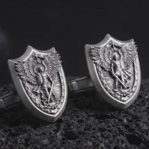 Puede incluir: Un par de gemelos de metal plateado, cada uno con forma de escudo. La cara de cada gemelo presenta un relieve detallado de una figura alada que sostiene una espada, de pie sobre una forma serpentina dentro del borde del escudo.