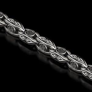 Può includere: Primo piano di una catena d'argento con un design ornato. Le maglie della catena sono di forma ovale con motivi dettagliati e uno sfondo scuro. La catena è realizzata in un materiale metallico lucido.