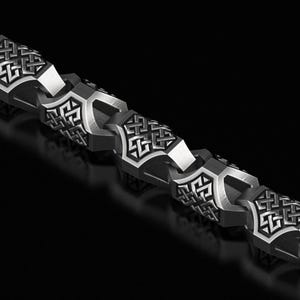 May include: A close-up shot of a silver chain bracelet. The bracelet has a geometric design with interconnected links. Each link has a detailed, symmetrical pattern. The links are a dark silver colour, set against a black background.