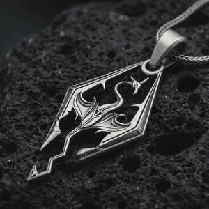 Oxidized Skyrim dragon pendant necklace on textured volcanic rock, close-up studio shot highlighting the angular dragon emblem, openwork design, and antique silver contrast.