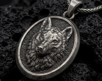 May include: A silver-toned oval pendant featuring a detailed, raised relief of a wolf head. The pendant has a dark, oxidised finish and is attached to a thick, woven silver-toned chain. The wolf design shows textured fur and pointed ears.