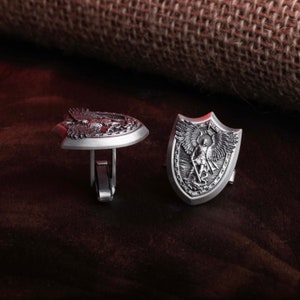 May include: A pair of silver cufflinks with detailed engravings. One cufflink features an eagle design, while the other depicts an angel with wings outstretched.