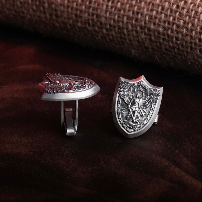 May include: A pair of silver cufflinks with detailed engravings. One cufflink features an eagle design, while the other depicts an angel with wings outstretched.