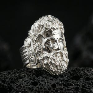 Polished silver Zeus ring featuring sculpted god portrait and refined facial expression, centered close-up jewelry photo on dark porous volcanic stone