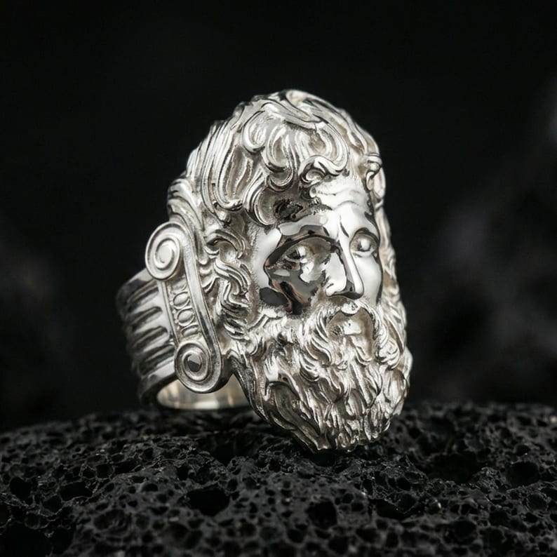 Polished silver Zeus ring featuring sculpted god portrait and refined facial expression, centered close-up jewelry photo on dark porous volcanic stone