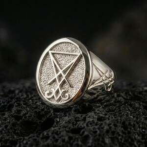Polished silver Lucifer sigil signet ring featuring a bold raised emblem and clean beveled edges, close-up on volcanic stone