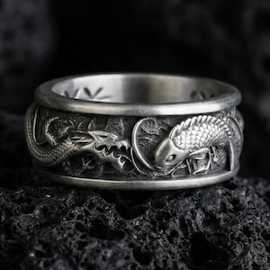 Oxidized sterling silver dragon and koi fish band ring on dark volcanic rock, Japanese-inspired relief details, premium macro product shot.