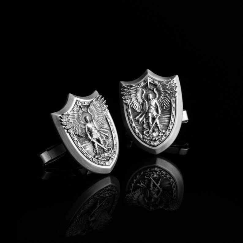 May include: A pair of silver cufflinks featuring a shield design with an angel holding a sword. The angel is depicted with wings and a halo.