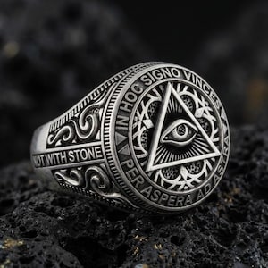 Oxidized sterling silver all-seeing eye triangle ring with Latin inscription and engraved scrollwork, macro on black volcanic rock