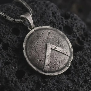 May include: A circular silver-toned pendant featuring a textured, weathered surface with a raised Lambda symbol in the centre. The pendant is attached to a silver-toned wheat chain, showcasing a distressed, oxidised finish throughout the metal.