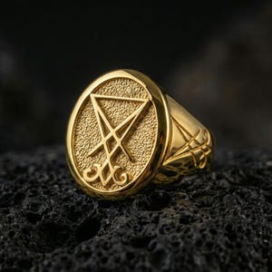 Gold plated Lucifer sigil signet ring with prominent raised occult symbol and dark textured background, macro on black volcanic rock