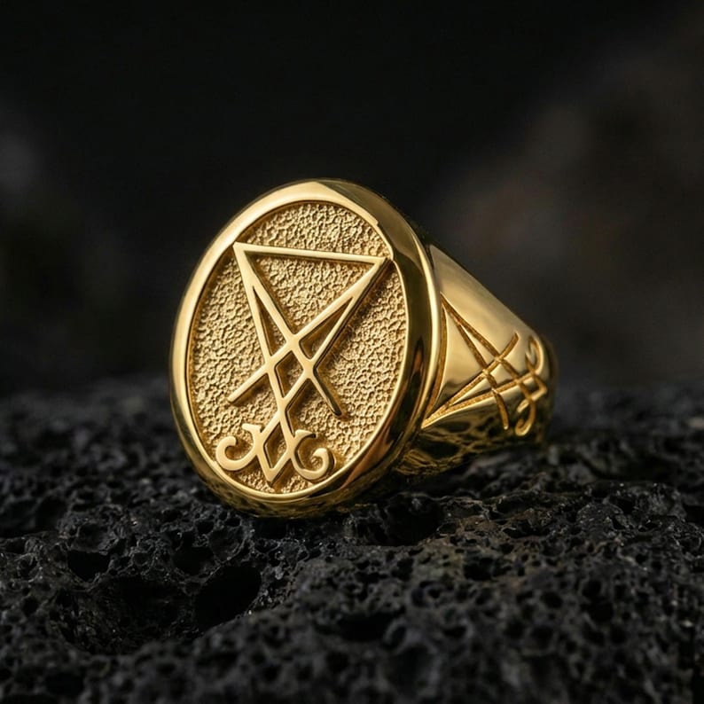 Gold plated Lucifer sigil signet ring with prominent raised occult symbol and dark textured background, macro on black volcanic rock